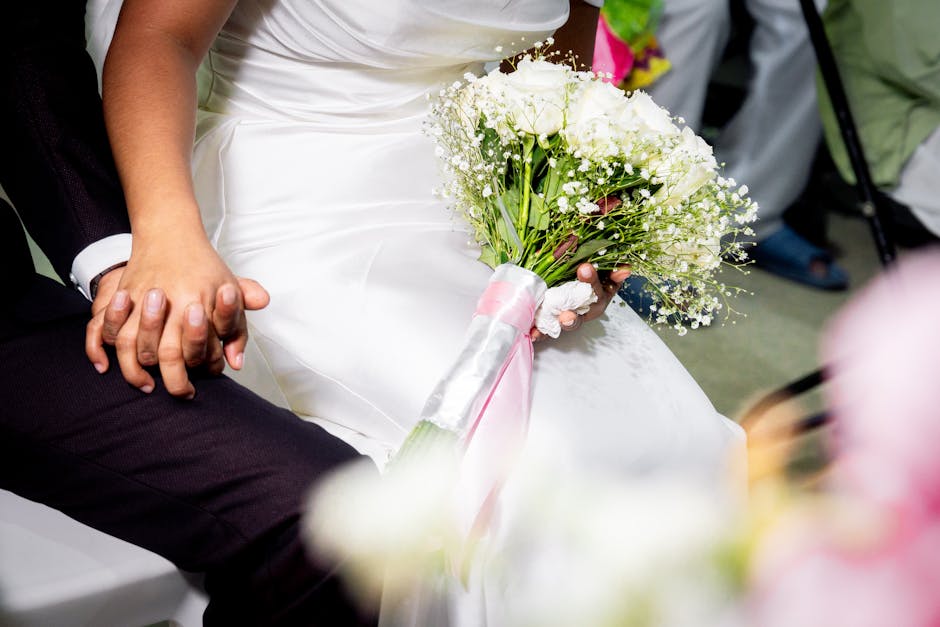 A close-up of a bridal bouquet placed on the bride's lap, featuring a collection of fresh white roses and delicate baby's breath flowers, wrapped in a white satin ribbon with pink accents. The bouquet