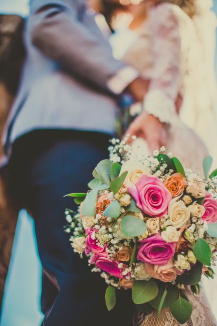 A close-up of a vibrant floral bouquet held by a bride during a wedding ceremony. The bouquet features an arrangement of fresh roses in shades of pink, peach, and cream, complemented by sprigs of baby