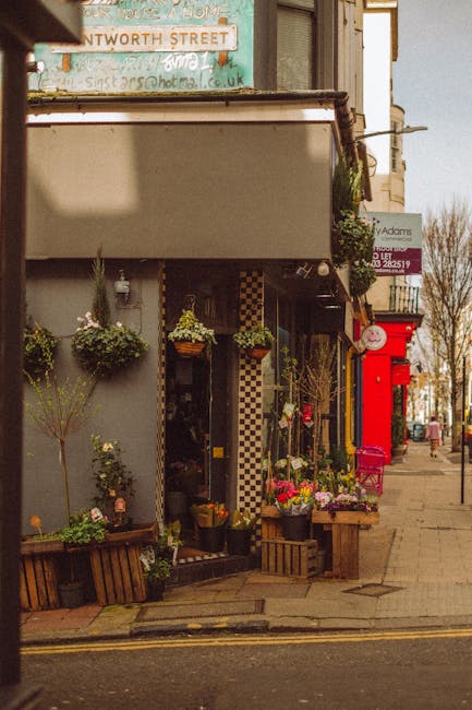 A floral display outside a florist shop on a street corner, showcasing a variety of fresh flowers including pink roses, white daisies, and purple irises arranged in bouquets and potted plants. The flo