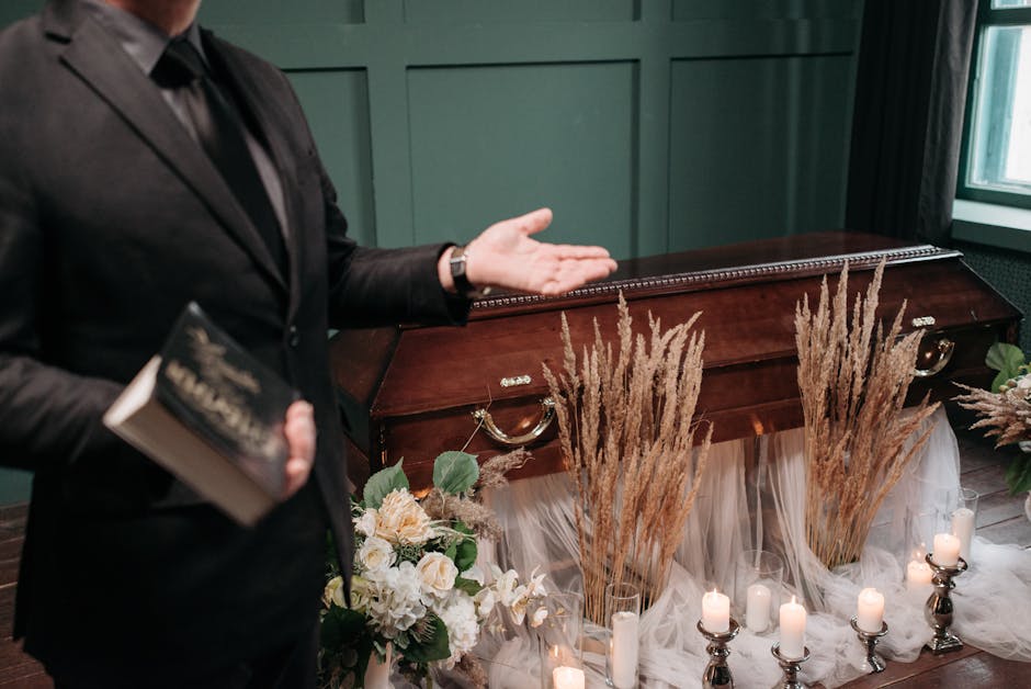 A floral arrangement for a funeral display featuring a mix of soft cream and white roses, beige dried pampas grass, and green foliage, arranged in a low display with white flowing tulle fabric and lit