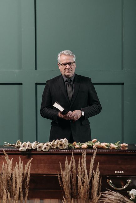 A middle-aged man with grey hair, glasses, and a serious expression, dressed in a black suit and tie, stands behind a wooden table decorated with a floral arrangement consisting of white and pale pink