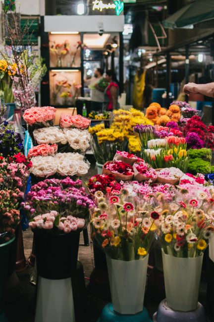 A vibrant flower market scene featuring an array of fresh floral arrangements and bouquets displayed on black and white stools and tables. The flowers include pink and white peonies, multicoloured tul