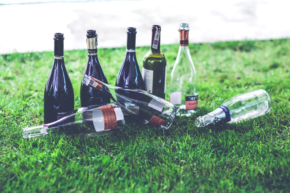 Several empty glass wine bottles, some with labels partially removed, are scattered on a patch of green grass outdoors. The bottles vary in shape and size, with a mix of dark and clear glass, and some