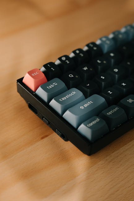 A close-up image of a compact mechanical keyboard placed on a wooden surface, featuring keycaps in pastel shades of pink, blue, and gray. The pink 'esc' key stands out at the top left corner, with blu