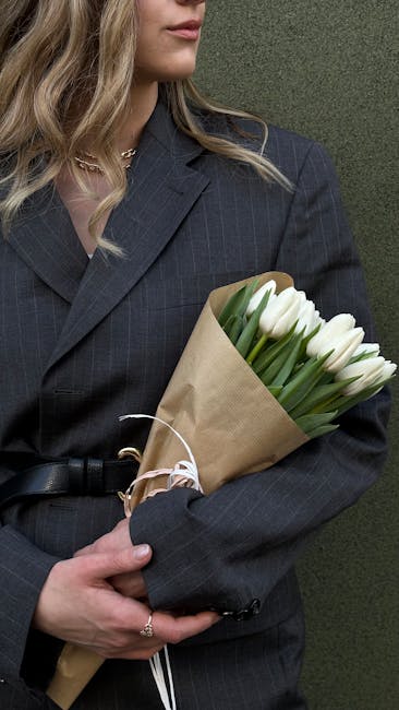 A woman dressed in a dark pinstripe blazer is holding a bouquet of fresh white tulips wrapped in brown kraft paper, secured with white ribbon. Her blonde hair cascades over her shoulder, and she is po